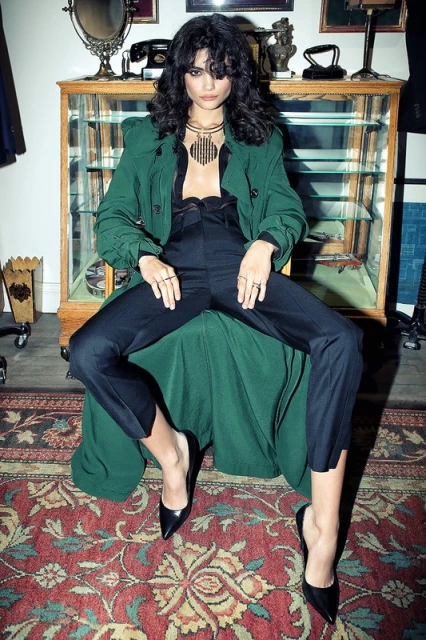 Fashionable adult female poses seated wearing a green trench coat, black top and pants, black heels, and gold jewelry indoors.