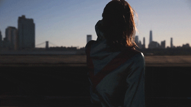 Woman in hooded jacket stands silhouetted against the sun, overlooking a city skyline with skyscrapers and a distant bridge at dusk.