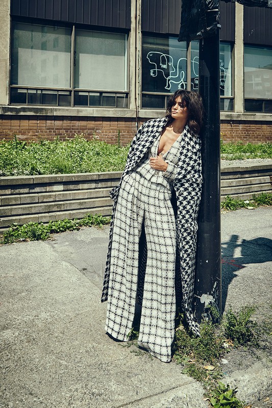 Fashionable woman in houndstooth coat poses against lamppost outside building. Style and elegance in an urban editorial photoshoot.