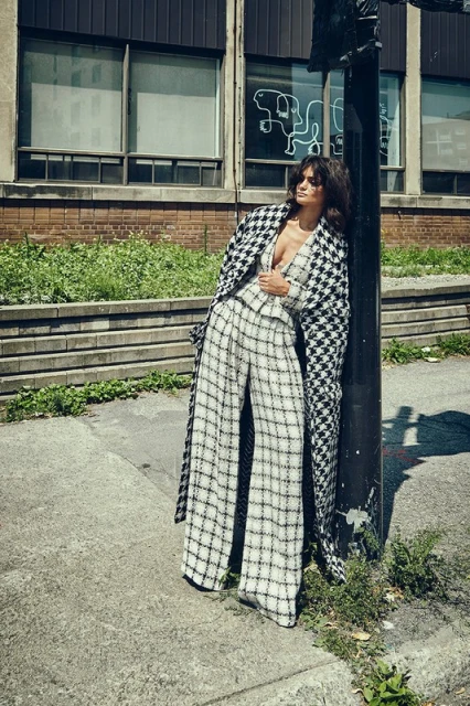 Fashionable woman in houndstooth coat poses against lamppost outside building. Style and elegance in an urban editorial photoshoot.