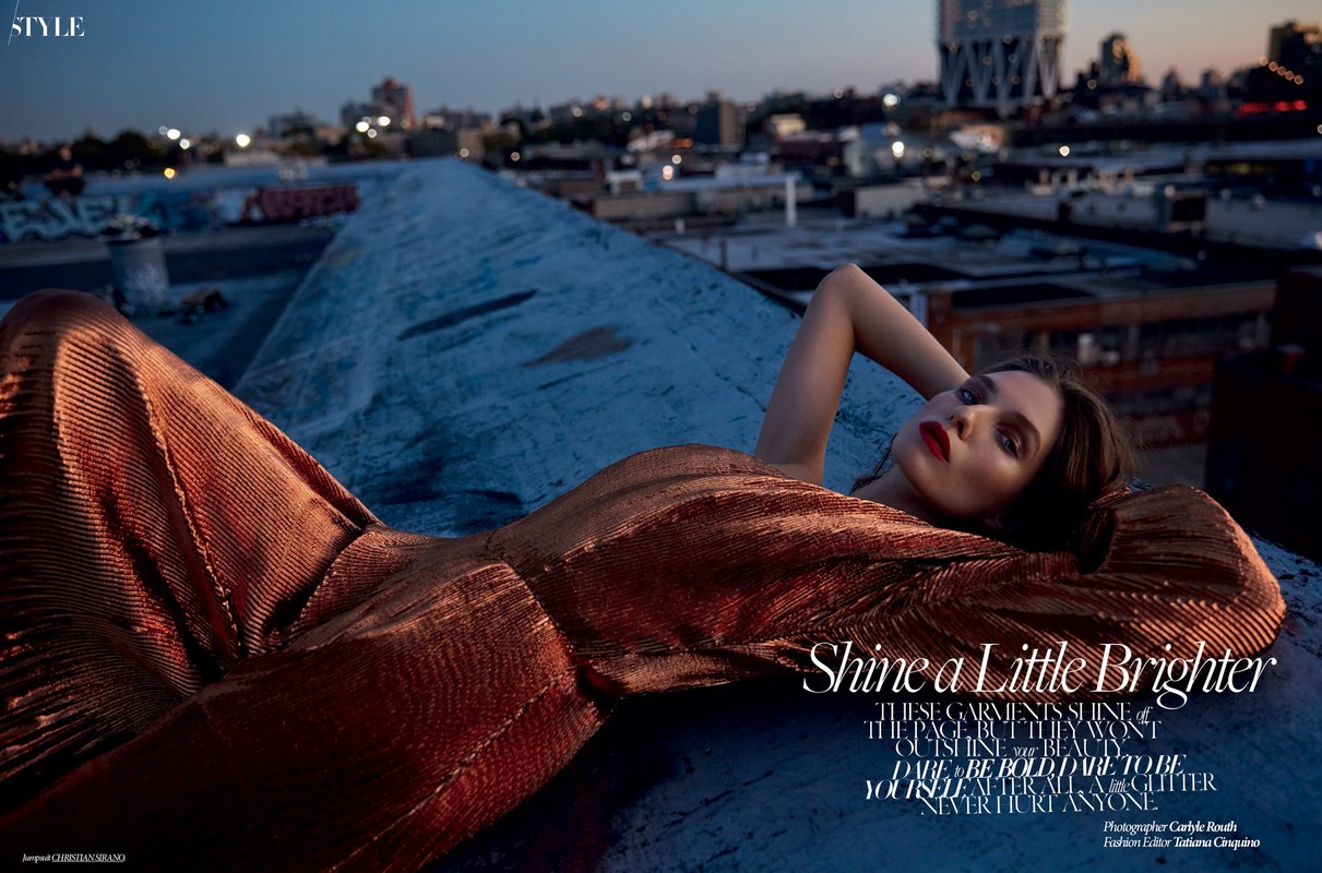 Fashion editorial shot of a reclining woman wearing a copper metallic jumpsuit on a rooftop against a city skyline at dusk.
