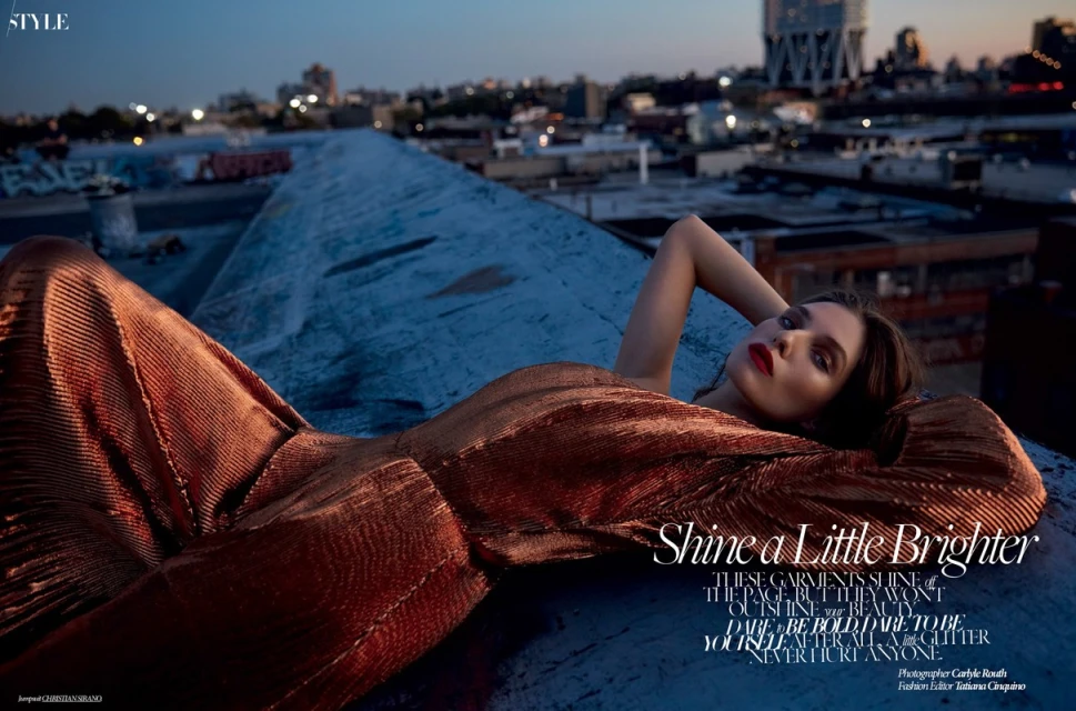 Fashion editorial shot of a reclining woman wearing a copper metallic jumpsuit on a rooftop against a city skyline at dusk.