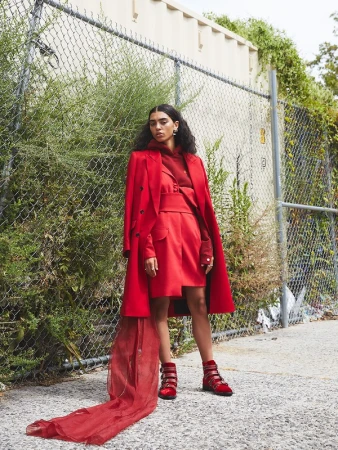 Model standing outdoors in a bright red outfit featuring a tailored coat, skirt, and buckled boots against a chain-link fence.