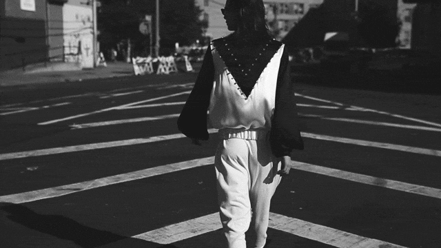 A lone adult wearing a black and white ensemble walks through an outdoor crosswalk on a sunny day; urban setting with buildings in the background.