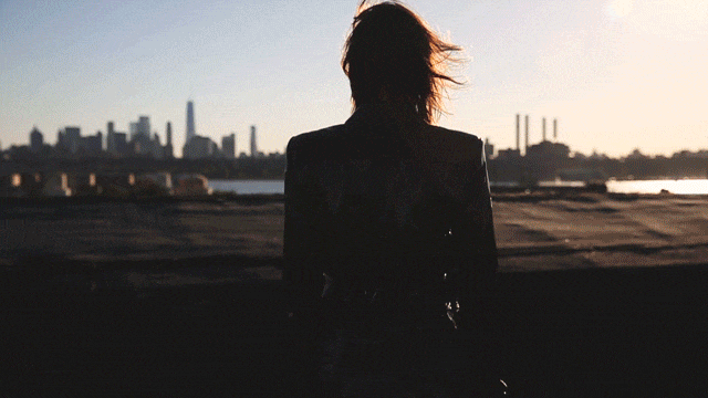 Person silhouetted against a city skyline with blowing hair. Warm lighting and shallow depth of field enhance the urban scene.