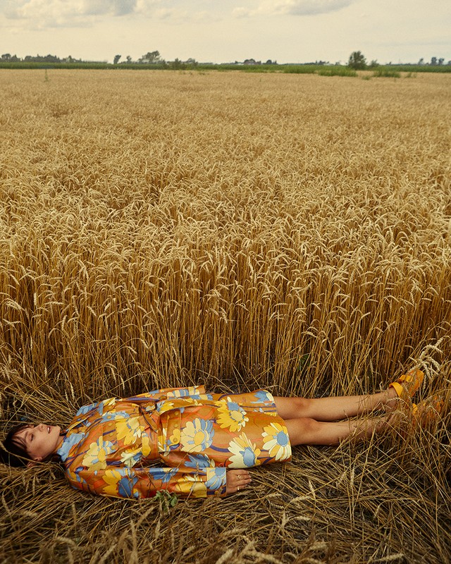 Woman lying down in a golden wheat field wearing a floral coat. The scene captures a peaceful moment in nature with clear sky.