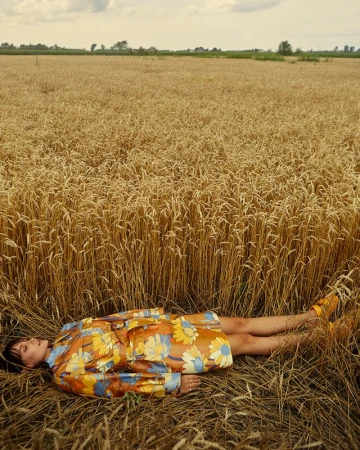 Woman lying down in a golden wheat field wearing a floral coat. The scene captures a peaceful moment in nature with clear sky.