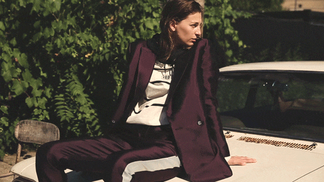 Adult female model wearing a burgundy blazer and sweatpants sitting on the hood of a cream-colored vintage car outdoors with green foliage.