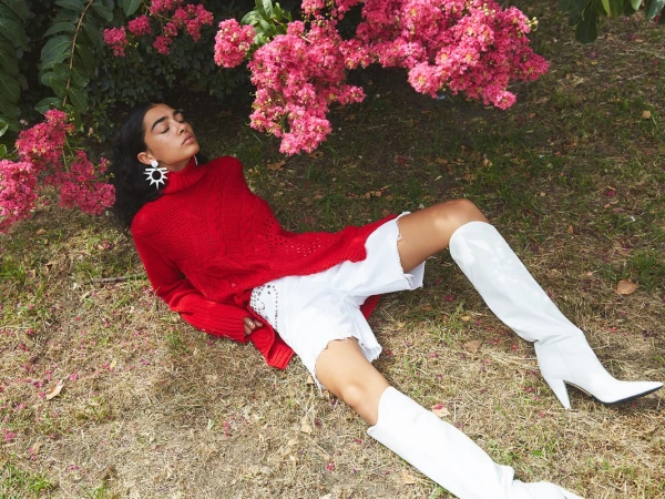Woman in red sweater and white boots reclines on grass under pink blossoms, outdoor fashion editorial.