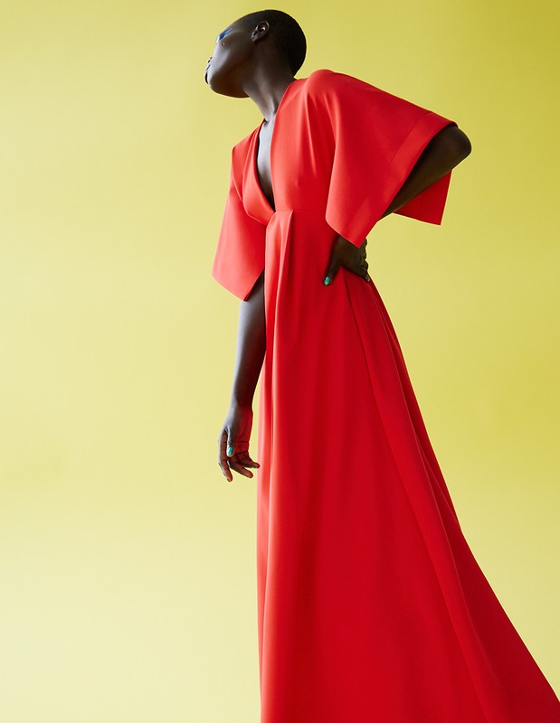 A woman with short hair poses in a flowing red dress against a bright yellow background, looking up and away with a hand on her hip.