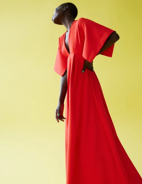 A woman with short hair poses in a flowing red dress against a bright yellow background, looking up and away with a hand on her hip.