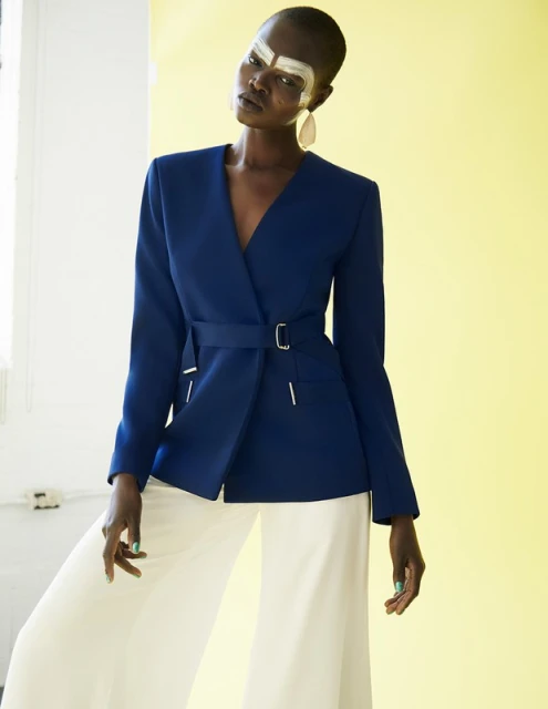 A dark-skinned woman in a blue belted jacket and white pants poses indoors against a yellow and white wall, showcasing makeup and fashion.