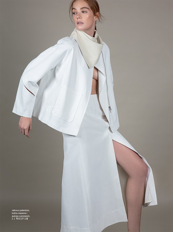 Fashion editorial shot: A red-haired model poses in a white jacket and long skirt with a thigh slit, set against a neutral backdrop.