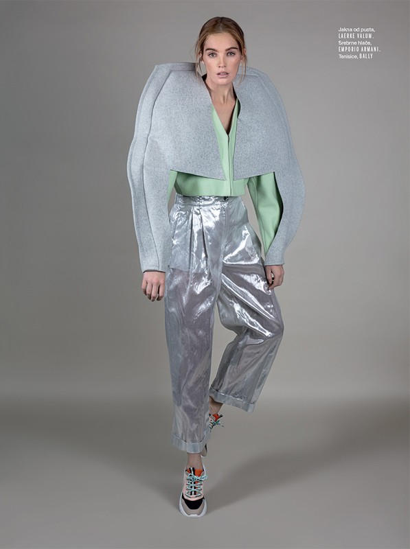 Fashion editorial shot of a model wearing a grey, structured bolero jacket, green top, and metallic silver pants with running shoes on grey backdrop.