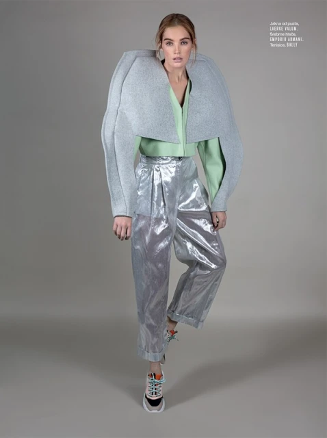 Fashion editorial shot of a model wearing a grey, structured bolero jacket, green top, and metallic silver pants with running shoes on grey backdrop.