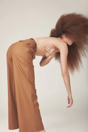 Model with voluminous curly brown hair wearing camel trousers bending over against a pale backdrop