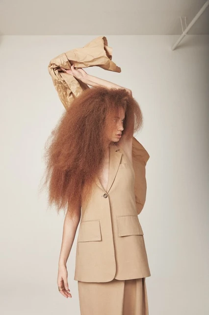 Fashionable woman with red hair in a beige vest and skirt holds a tan paper bag above her head in a studio setting with soft lighting.