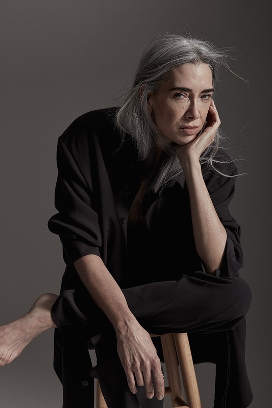 Elegant mature woman with long grey hair poses in black on a wooden stool in a studio setting with subdued lighting.