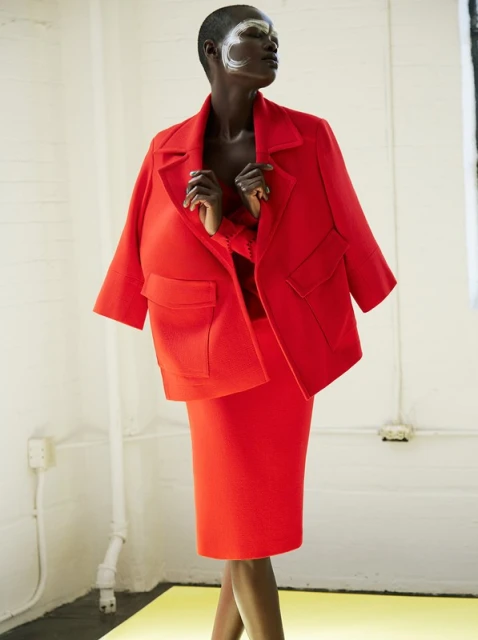 Dark-skinned woman in a red skirt suit with white eye makeup poses in a minimalist white studio with a bright yellow floor.