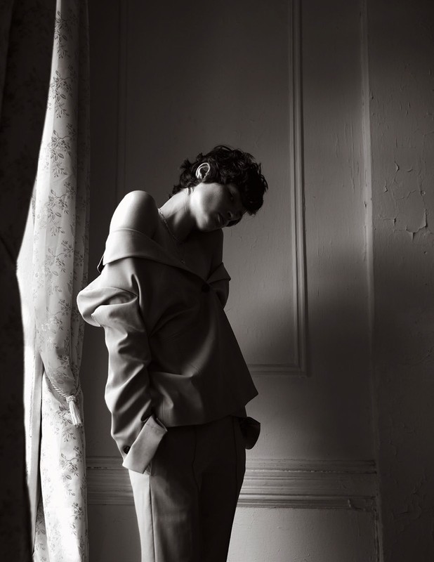 Black and white portrait of an adult female in an off-the-shoulder top and tailored pants, posed indoors near a window with a curtain.