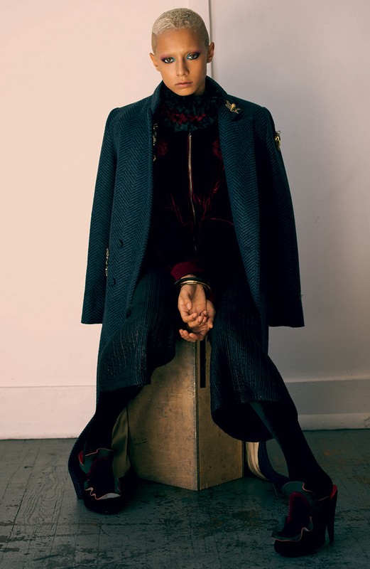 A woman with bleached blonde hair poses sitting on a cube in an indoor studio wearing an overcoat, velvet top, pleated pants, and platform shoes.