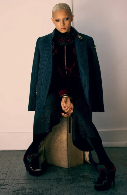 A woman with bleached blonde hair poses sitting on a cube in an indoor studio wearing an overcoat, velvet top, pleated pants, and platform shoes.