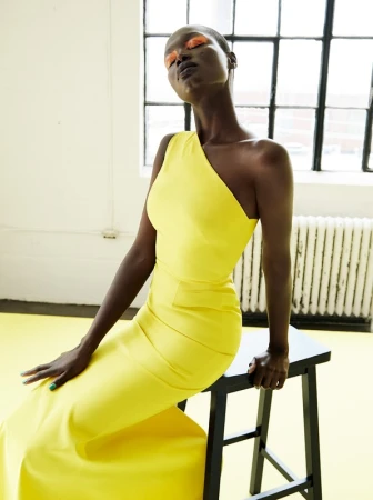 A dark-skinned woman poses indoors wearing a yellow one-shoulder dress, seated on a stool in front of a bright window.