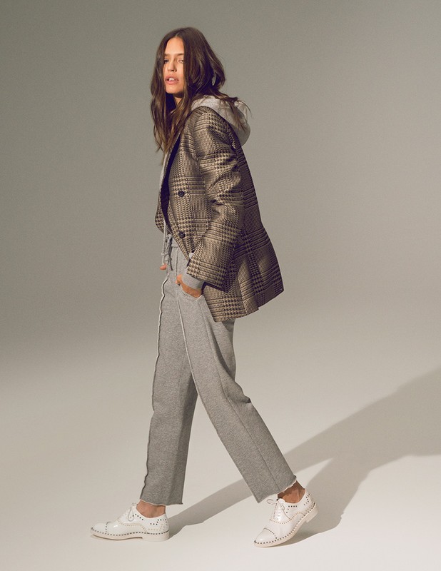 Full-length studio shot of a woman in a gray tracksuit, hooded sweatshirt, a plaid blazer, and white brogues walking and looking left.