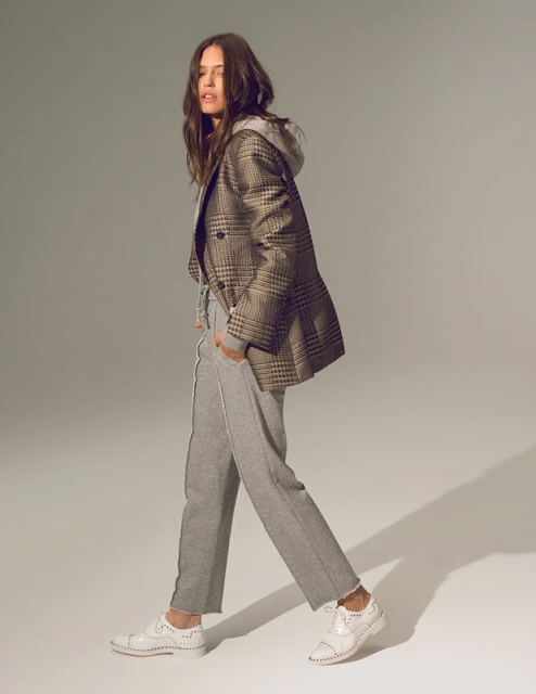 Full-length studio shot of a woman in a gray tracksuit, hooded sweatshirt, a plaid blazer, and white brogues walking and looking left.