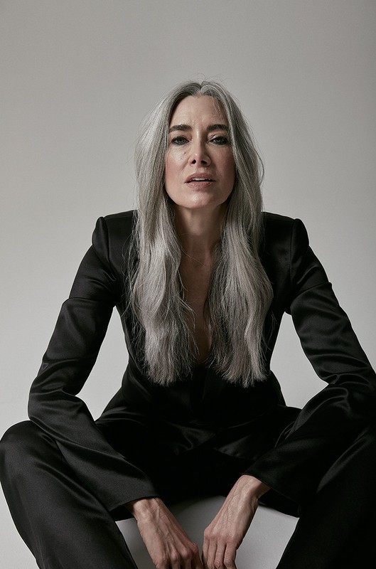 Portrait of a mature woman with long gray hair wearing a black suit sitting in a photo studio against a white background looking at the camera.