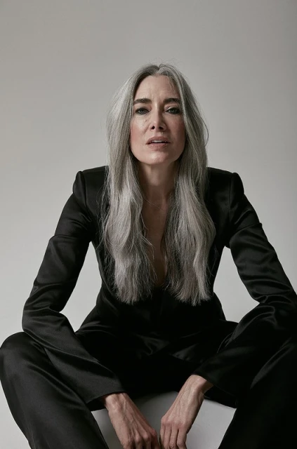 Portrait of a mature woman with long gray hair wearing a black suit sitting in a photo studio against a white background looking at the camera.