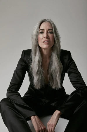 Portrait of a mature woman with long gray hair wearing a black suit sitting in a photo studio against a white background looking at the camera.