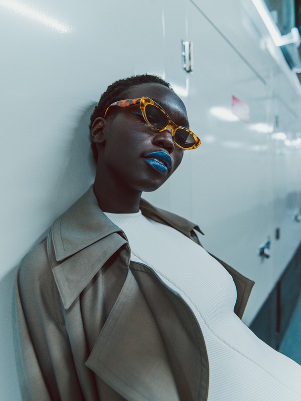 Stylish woman with dark skin and blue lipstick wears retro sunglasses and a trench coat, posing against a sleek, glossy white wall.