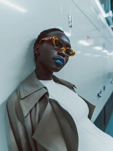 Stylish woman with dark skin and blue lipstick wears retro sunglasses and a trench coat, posing against a sleek, glossy white wall.