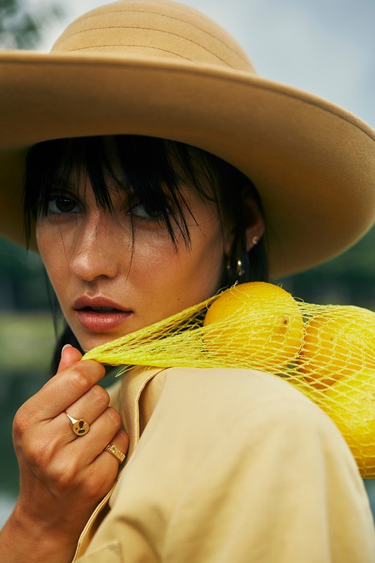 Woman wearing a wide-brimmed hat with a net bag of lemons draped over her shoulder, posing outdoors with neutral clothing and jewelry.