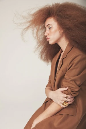 Profile portrait of a woman with voluminous brown hair wearing a brown blazer against a plain white background