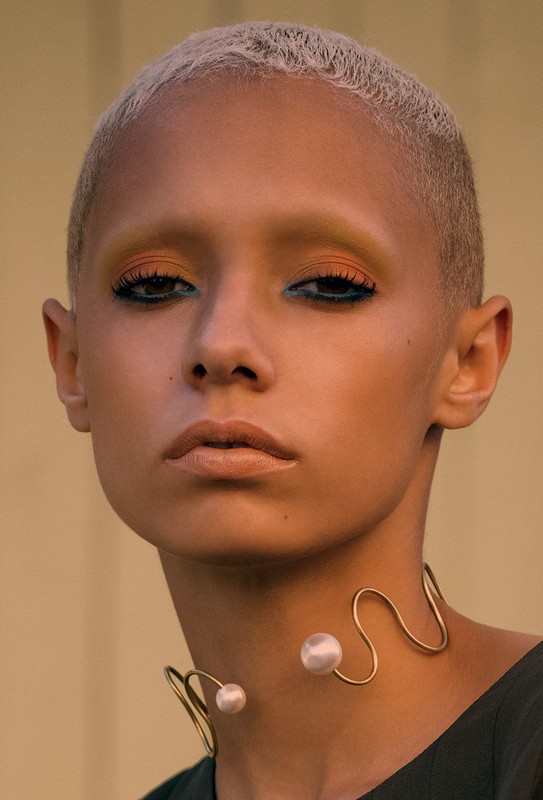 Close-up portrait of a woman with a short blonde haircut, wearing bold eye makeup and a distinctive gold and pearl statement necklace.