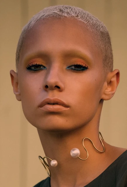 Close-up portrait of a woman with a short blonde haircut, wearing bold eye makeup and a distinctive gold and pearl statement necklace.