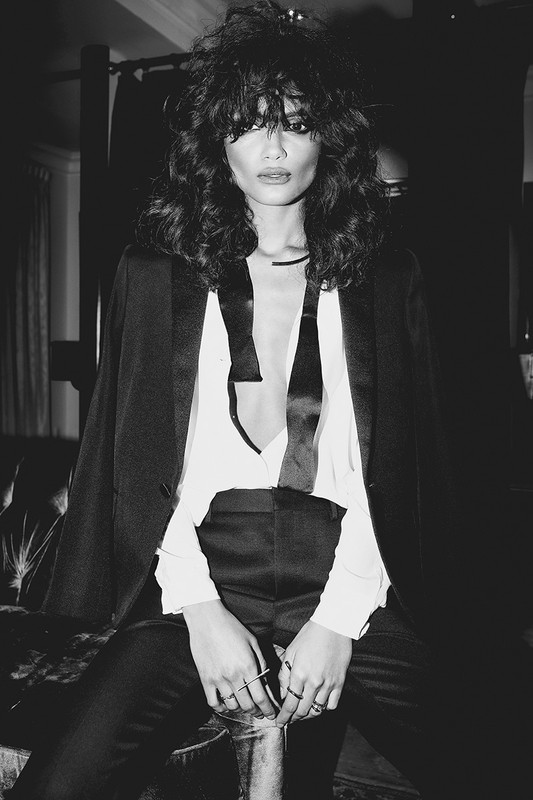 A black and white portrait of a stylish adult woman with wavy dark hair, wearing a black suit jacket over a white top, sitting indoors.