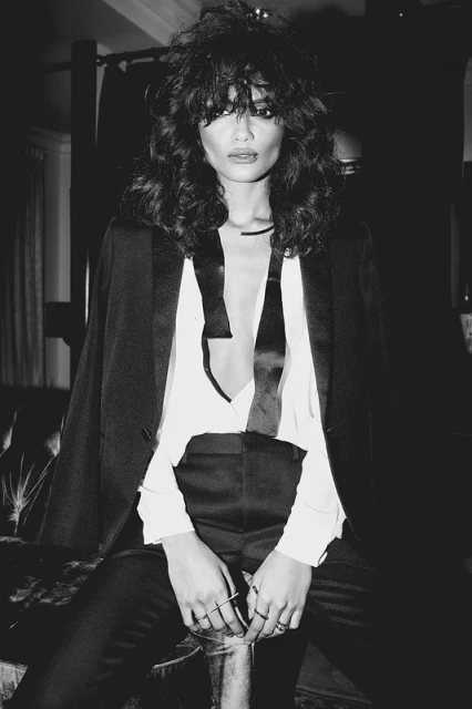 A black and white portrait of a stylish adult woman with wavy dark hair, wearing a black suit jacket over a white top, sitting indoors.