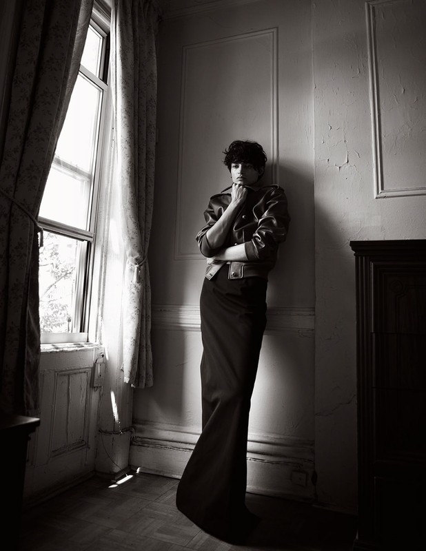 Monochromatic portrait of a person leaning against a wall in a room with a window, wearing a jacket and long skirt or dress.