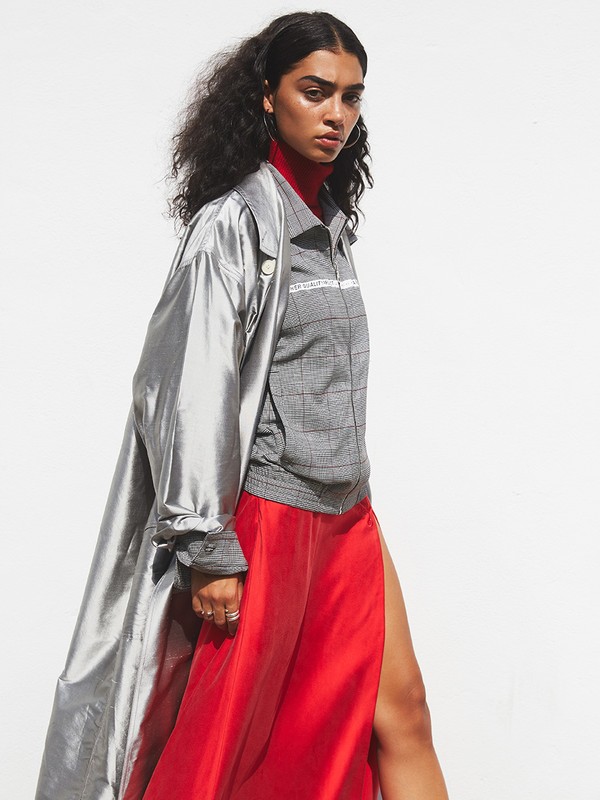 Woman in a gray plaid top and red skirt with a silver jacket poses in a fashion shot against a white backdrop in a studio.