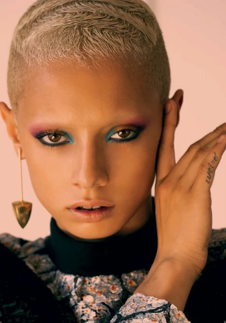 Close-up portrait of a young adult woman with short blonde hair, colorful eye makeup, an earring, and a patterned top.