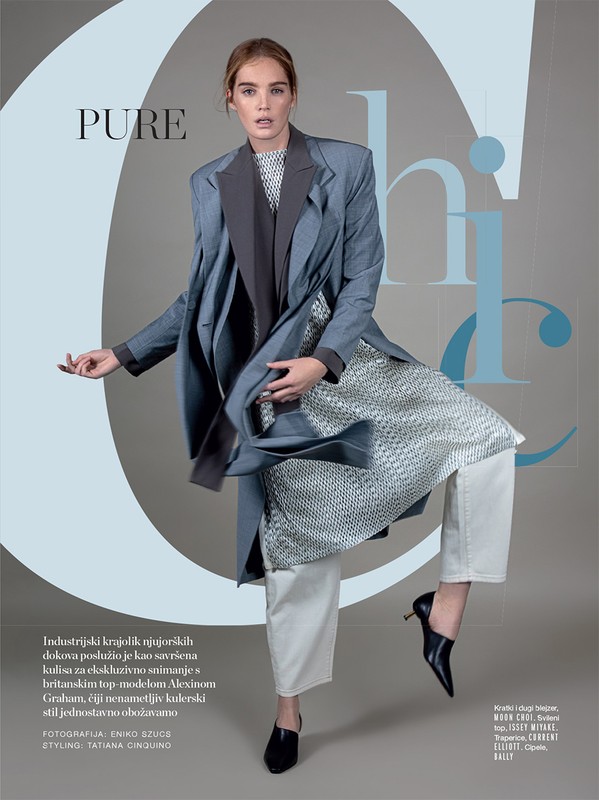 Fashion editorial shot of a young adult woman in an oversized coat, white pants, and black heeled shoes in a studio setting.