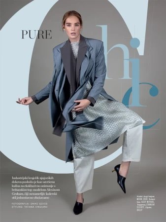 Fashion editorial shot of a young adult woman in an oversized coat, white pants, and black heeled shoes in a studio setting.