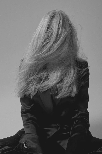 Back view of a woman with long blonde hair and a black satin jacket against a grey background. The hair is covering her face.