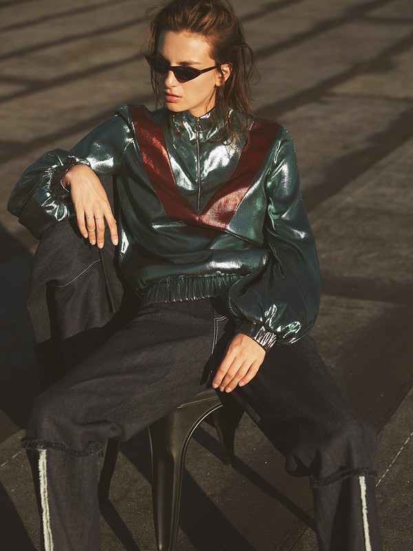 A woman in a shiny jacket and dark jeans sits on a stool, looking to the side against a concrete surface in an outdoor setting.