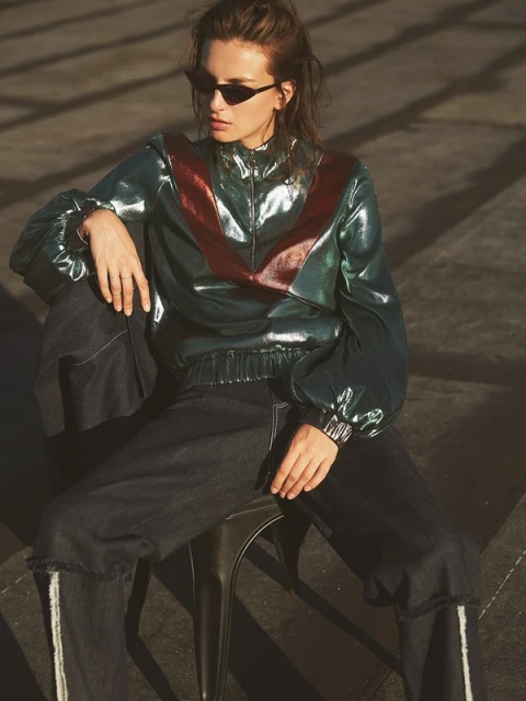 A woman in a shiny jacket and dark jeans sits on a stool, looking to the side against a concrete surface in an outdoor setting.