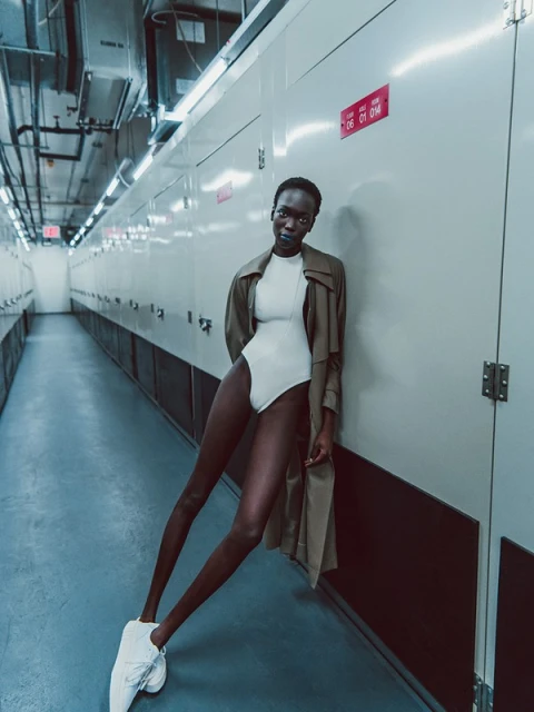 Adult woman strikes a pose in a stylish bodysuit and trench coat while standing inside a long hallway with locker-style storage units.