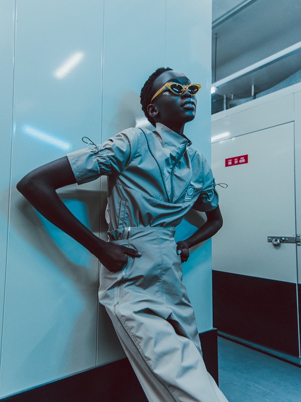 Stylish woman in beige two-piece outfit and yellow sunglasses poses against an indoor white wall with a modern, fashionable look.